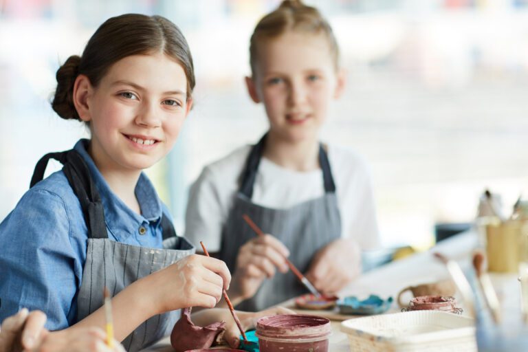 Little schoolgirl with paintbrush and her classmate sitting by table at lesson and creating clay items